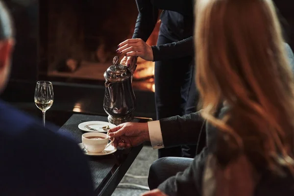 女服务员在白杯中倒入热茶 朋友们坐在有漂亮壁炉的餐厅里 — 图库照片
