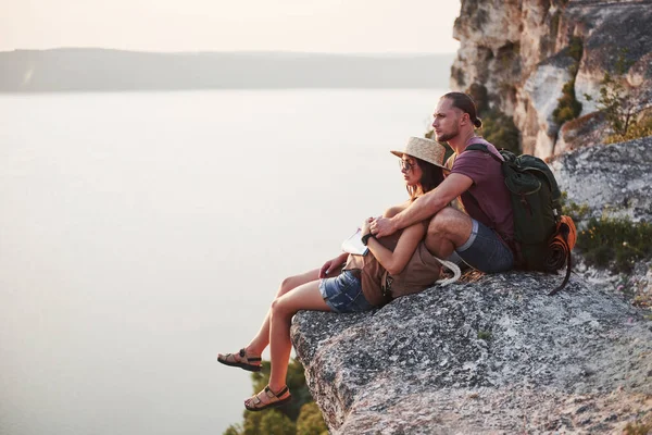 岩の山の上に座って川や湖の海岸の景色を楽しむバックパック付きの巨大なカップル 山や海岸を旅し 自由とアクティブなライフスタイルのコンセプト — ストック写真