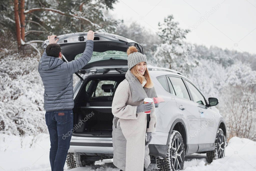 Woman with drink in the hands is smiling when guy closing trunk of the modern silver colored car. Couple have arrived to their destination of beautiful winter forest.