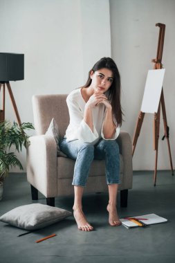Easel arkada. Beyaz duvarları ve pencereden gelen gün ışığıyla odada genç bir esmer..