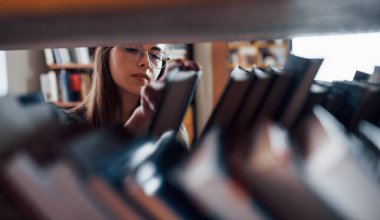 Rafların arasından bak. Kütüphanede kitaplarla dolu bir kız öğrenci var. Eğitim kavramı.