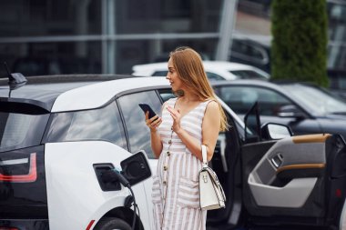 Şarj beklerken telefonu kullanmak. Gündüz vakti istasyonda bir kadın. Yepyeni bir araç..