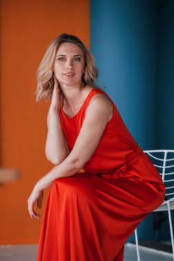 Kutlama geliyor. Şenlikli kırmızı elbiseli güzel sarışın turuncu ve mavi renkli duvarları ve beyaz sandalyesi olan odada..