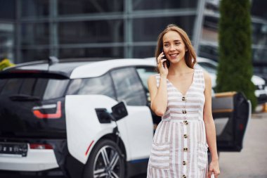 Şarj beklerken telefonu kullanmak. Gündüz vakti istasyonda bir kadın. Yepyeni bir araç..