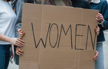 Elinde kocaman bir poster var. Bir grup feminist kadın dışarıda hakları için protesto yapıyor..