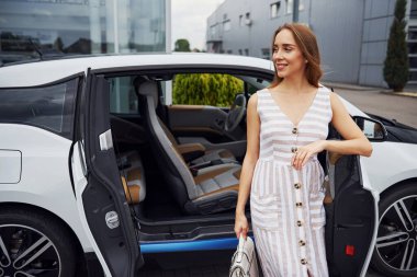 Elinde torbayla. Sarı saçlı kadın sokakta beyaz arabasının yanında duruyor..