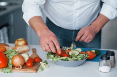 Küçük domatesleri salataya koymak. Beyaz gömlekli adam mutfakta sebze kullanarak yemek hazırlıyor..