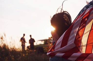 İnsanların önünde bir kız. Arkadaşlarının hafta sonları açık havada yeşil arabalarının yanında Amerikan bayrağı taşıyorlar..