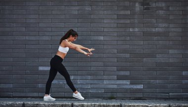 Genç, sportif esmer, ince vücutlu, şehirde gündüz vakti tuğla duvara yaslanmış..