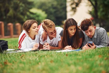 Kağıda yazıyorum. Ön manzara. Gündüz vakti yeşil çimlerde günlük giysiler içinde bir grup genç öğrenci..