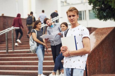 Kameraya poz veren adam. Gündüz vakti üniversitenin yakınlarında günlük giysiler içinde bir grup genç öğrenci.