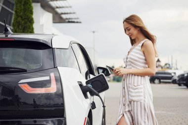 Beklerken akıllı telefon kullanmak. Elektrikli arabalardaki kadın gündüz şarj istasyonunda. Yepyeni bir araç..