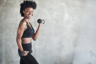 Duvara karşı çalışıyor. Elinde dambıllarla gri arka planda duran genç bir kadının stüdyo çekimi..