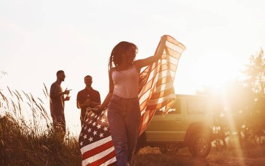 Kız öne doğru koşuyor. Arkadaşlarının hafta sonları açık havada yeşil arabalarının yanında Amerikan bayrağı taşıyorlar..