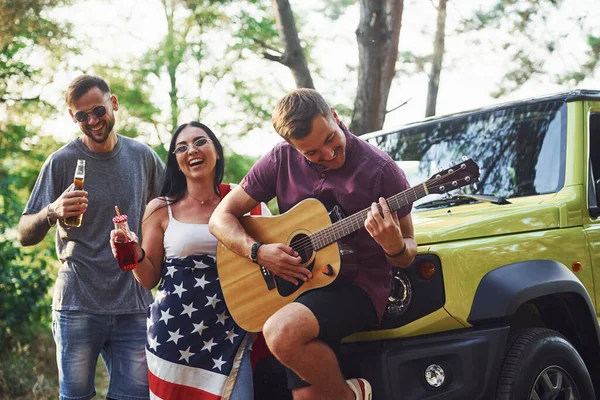 Musician plays a song on guitar. Friends have nice weekend outdoors near theirs green car with USA flag.