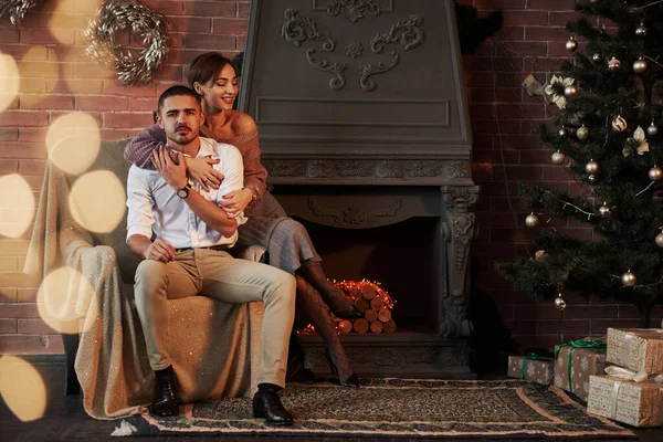 偉大な暖炉の近く 豪華な部屋で抱きかかえている椅子の上の美しい若いカップル休日の木とガーランド — ストック写真