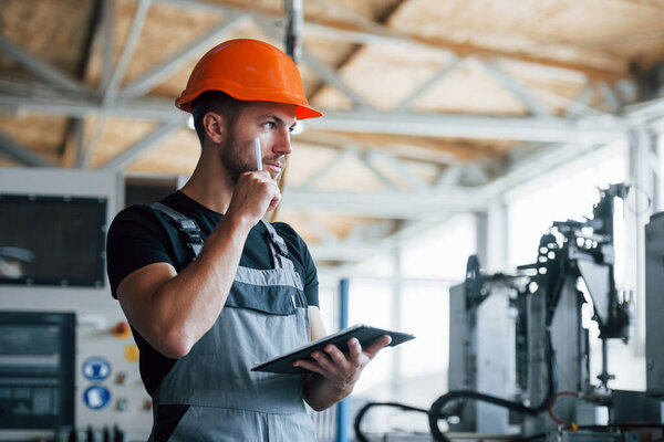 With notepad in hands. Industrial worker indoors in factory. Young technician with orange hard hat.