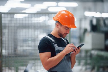 Akıllı telefon ellerde. Fabrikada sanayi işçisi. Turuncu şapkalı genç teknisyen..