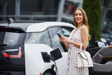 Şarj beklerken telefonu kullanmak. Gündüz vakti istasyonda bir kadın. Yepyeni bir araç..