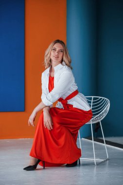 Ve çok da rahat. Şenlikli kırmızı elbiseli güzel sarışın turuncu ve mavi renkli duvarları ve beyaz sandalyesi olan odada..