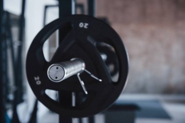 Halterin gümüş renkli kısmına odaklanmış. Spor salonundaki metal standında gündüz vakti siyah halter. Etrafta kimse yok..