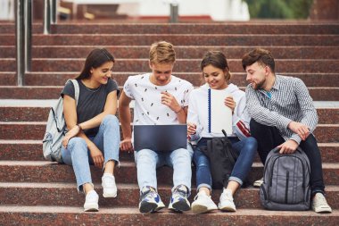 Dizüstü bilgisayarla. Gündüz vakti üniversitenin yakınlarında günlük giysiler içinde bir grup genç öğrenci.