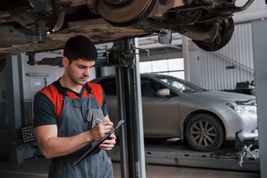 Asılan aracın altında. Üniformalı atölyedeki adam bozuk arabayı tamir etmek için not defteri kullanıyor..