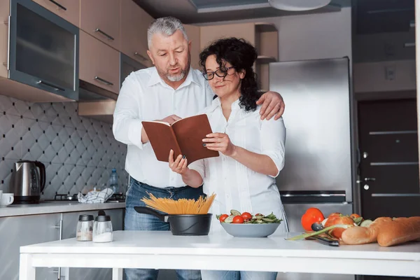 レシピの秘密の本だ 台所で野菜を使った料理を作る白いシャツを着た男と妻 — ストック写真