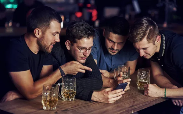 Funny content on smartphone. Three friends resting in the pub with beer in hands. Having conversation.