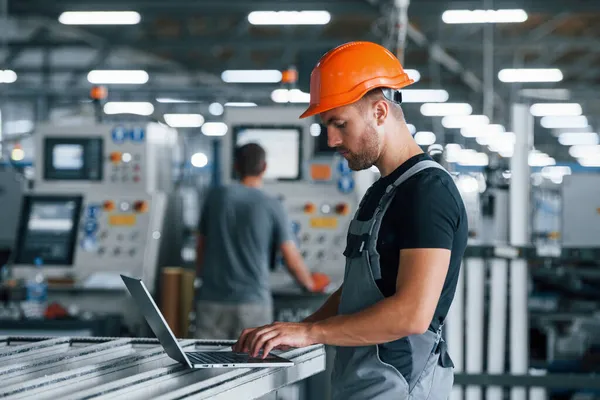 Modern laptop. Klavyede yazıyorum. Fabrikada sanayi işçisi. Turuncu şapkalı genç teknisyen..