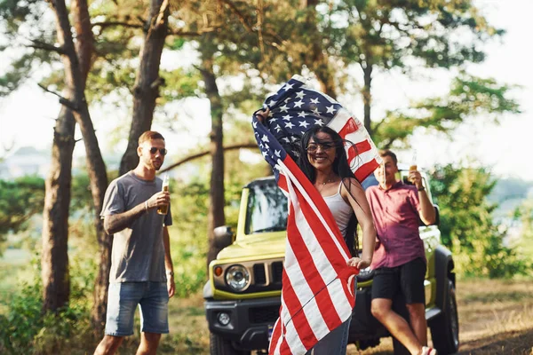In the forest. Friends have nice weekend outdoors near theirs green car with USA flag.