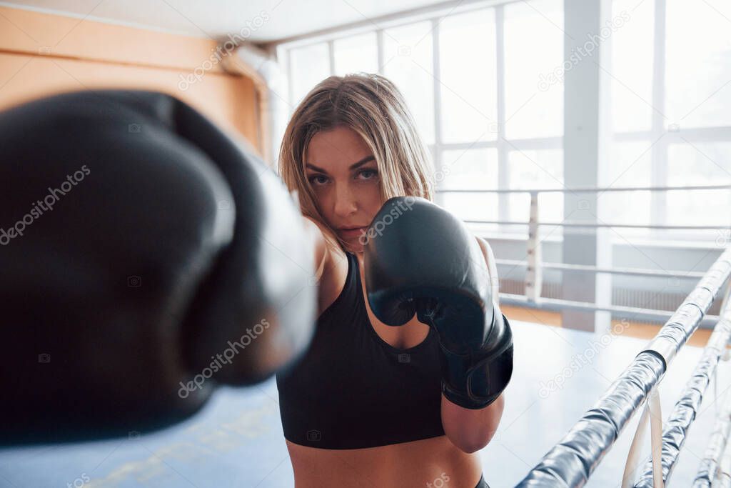 Punch in the face. Female sportswoman training in the boxing ring. In black colored clothes.