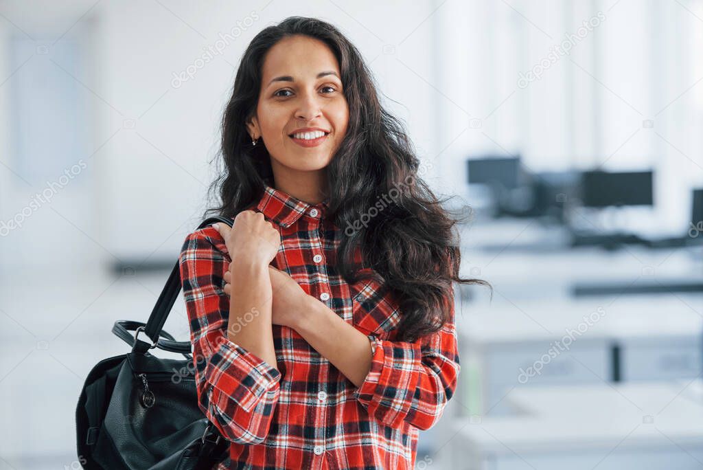 In casual clothes. Portrait of attractive young woman standing in the office with black bag.