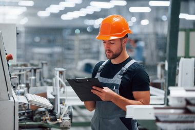 Ekipmanlara bakar ve notlar alır. Fabrikada sanayi işçisi. Turuncu şapkalı genç teknisyen..