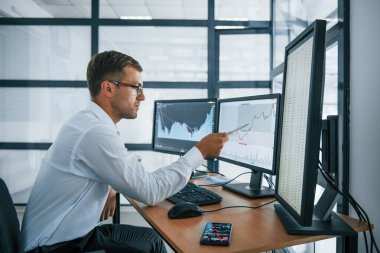 Grafikler analiz ediliyor. Resmi giyinmiş genç iş adamı birden fazla ekranla ofiste. Değişim ve para kavramı.