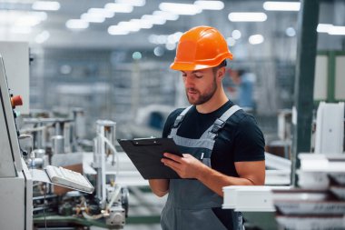 Ekipmanlara bakar ve notlar alır. Fabrikada sanayi işçisi. Turuncu şapkalı genç teknisyen..