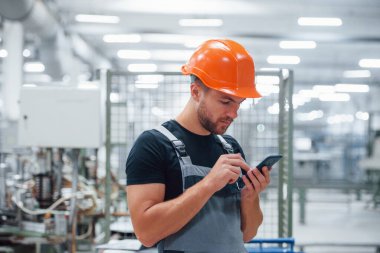 Akıllı telefon ellerde. Fabrikada sanayi işçisi. Turuncu şapkalı genç teknisyen..