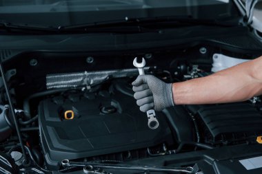 Dükkanda. Eldiven giymiş bir adam bozuk bir otomobilin önünde ingiliz anahtarı tutuyor..