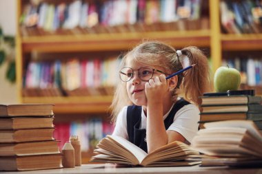 Saçları örgülü şirin küçük kız kütüphanede. Kitaplardaki elma..