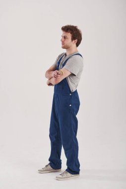 Kollarını kavuşturarak. Mavi üniformalı adam stüdyoda beyaz arka plana karşı duruyor..