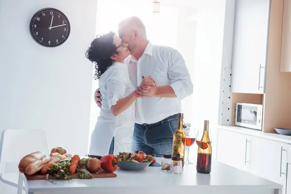 サイドビュー シニア男性と彼の妻で白いシャツ持っていますロマンチックなディナーオンザキッチン — ストック写真
