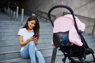 Merdivenlerde oturuyor, ellerinde akıllı telefon ve yanında pembe bebek arabasıyla. Güzel genç bir kadın hafta sonunu şehirde geçirir..