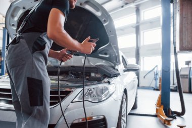 Kırmızı atlama kablolarını tutuyor. Gri üniformalı adam içeride beyaz otomobil tamir ediyor..