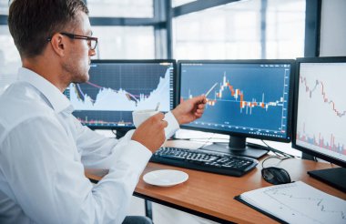 Bir bardak içki tutuyor. Bilgi analiz ediliyor. Resmi giyinmiş genç iş adamı birden fazla ekranla ofiste. Değişim ve para kavramı.