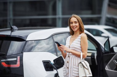Şarj beklerken telefonu kullanmak. Gündüz vakti istasyonda bir kadın. Yepyeni bir araç..
