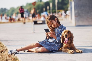 Yerde oturuyordu. Akıllı telefon ellerindeyken. Şirin küçük kız, güneşli bir günde köpeğiyle dışarı çıkar..
