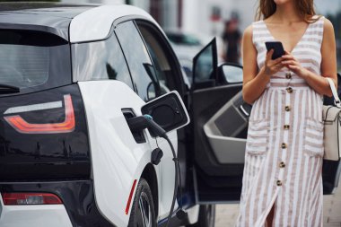 Modern kablosuz teknolojiler. Telefonla. Elektrikli arabalardaki kadın gündüz şarj istasyonunda. Yepyeni bir araç..