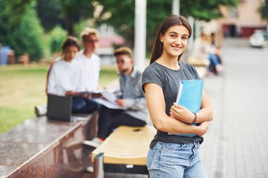 Elinde mavi klasör olan kız. Gündüz vakti şehirde günlük kıyafetler giyen bir grup genç öğrenci..