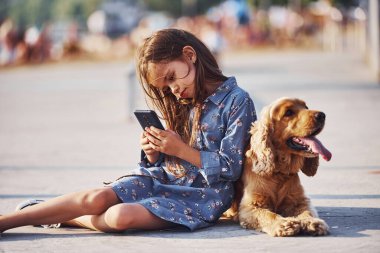Yerde oturuyordu. Akıllı telefon ellerindeyken. Şirin küçük kız, güneşli bir günde köpeğiyle dışarı çıkar..
