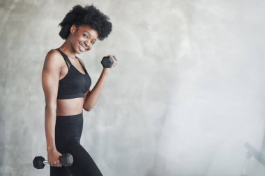 Güzel vücut. Elinde dambıllarla gri arka planda duran genç bir kadının stüdyo çekimi..